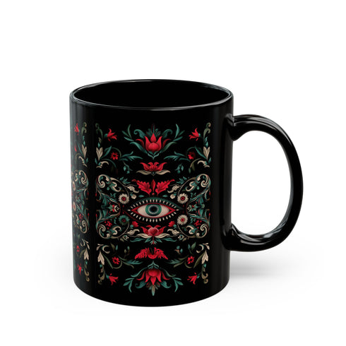 A black ceramic mug featuring a symmetrical folk art design with a central mystic eye motif in turquoise and coral. The design includes ornate scrollwork, red tulip-like flowers, sage green leaves, and cream flourishes arranged in a decorative pattern. The mug has a glossy finish with a C-shaped handle.